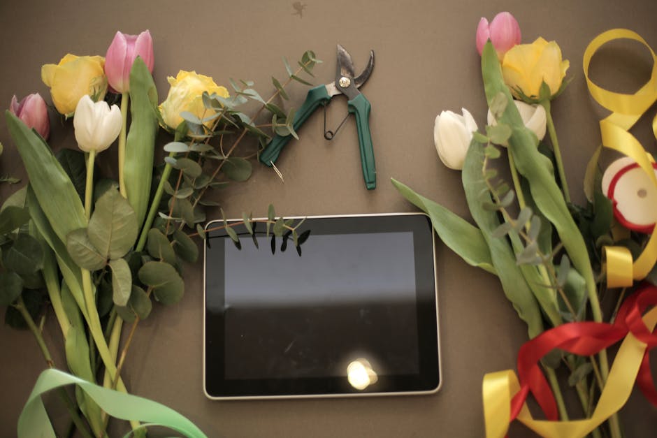 A flat lay arrangement of fresh flowers, including pink and white tulips and yellow roses, decorated with sprigs of green eucalyptus. The flowers are partially wrapped with colorful ribbons in red, ye