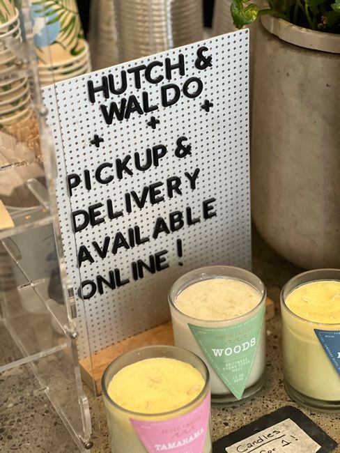 Close-up of a white pegboard sign with black raised lettering that reads 'Hutch & Waldó pickup & delivery available online!', positioned next to a large, round, textured ceramic pot containing a gree